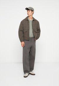 Brun- og sortternet jakke, grøn sweater, grå brede bukser og grå kasket. Lysebrune sneakers med hvide snørebånd og brune accenter.