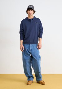 Ναυτικό φούτερ με γιακά και λογότυπο, σε συνδυασμό με oversized ανοιχτό μπλε τζιν παντελόνι. Καφέ σουέτ παπούτσια ολοκληρώνουν την εμφάνιση.