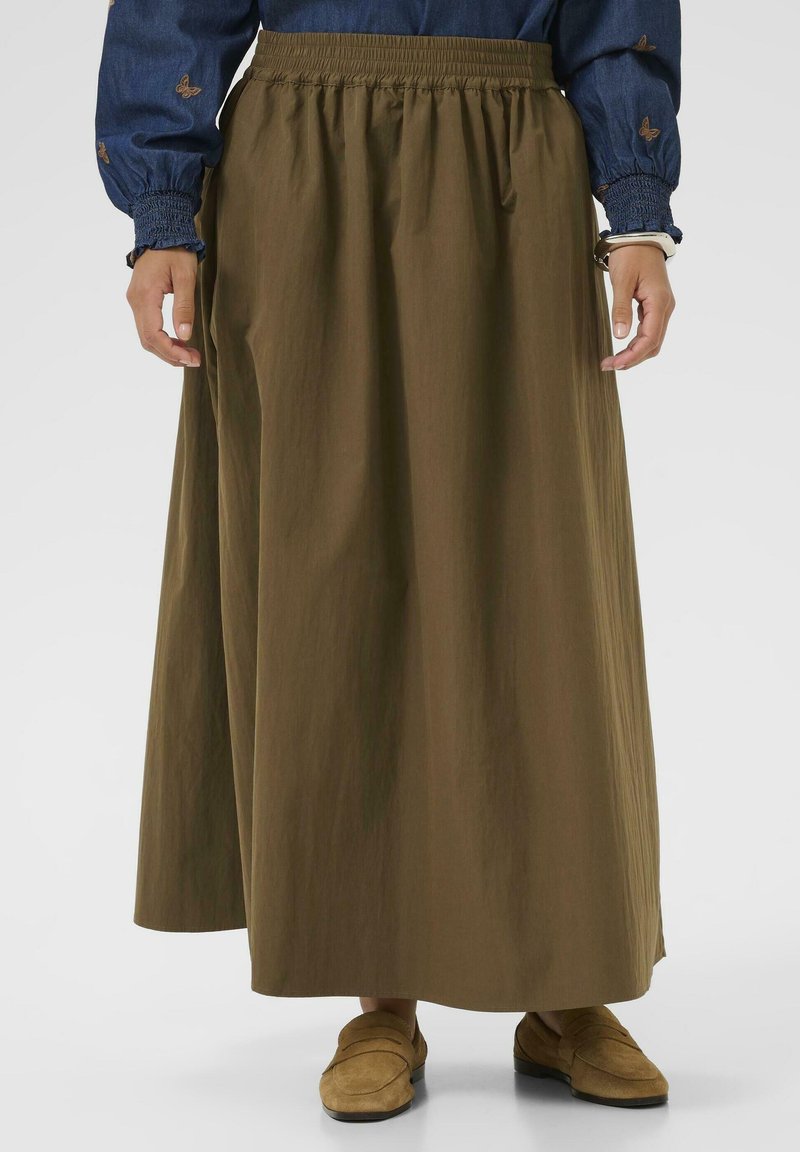 Kvindelig iført en lang brun nederdel, en blå bluse med sommerfuglebroderi og brune loafers, stående mod en ensfarvet baggrund.