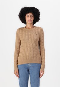 Pull en tricot beige avec motif en cable, encolure et poignets côtelés. Présente un petit logo sur la poitrine gauche. Associé à un jean bleu.