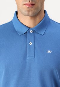 Lyseblå polo-shirt med tekstureret stof, to knapper ved kraven og et lille logo på venstre side. Kraven er struktureret.
