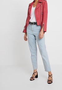 Czerwona zamszowa kurtka motocyklowa z srebrnymi zamkami, biała koszulka z dekoltem w serek, jasnoniebieskie dżinsy i czarne sandały na obcasie z paskami.