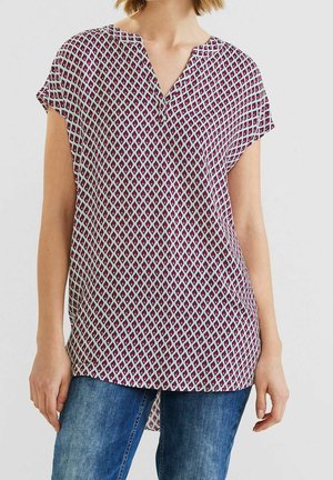 Femme portant un chemisier à manches courtes et col en V avec un motif losange rose et noir, associé à un jean bleu, debout devant un fond uni.