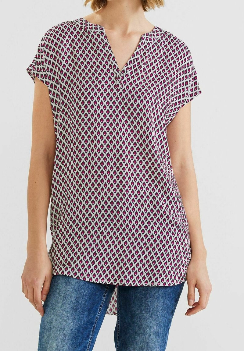 Femme portant un chemisier à manches courtes et col en V avec un motif losange rose et noir, associé à un jean bleu, debout devant un fond uni.