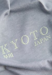 Textil gris con el texto grabado "KYOTO" en verde claro y "JAPÓN" debajo, con una textura de ribete horizontal evidente en la tela.