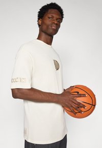 Kremfarget ribbestrikket kortermet skjorte med brodert "Celtics"-logo, parret med teksturerte svarte shorts og en oransje basketball.
