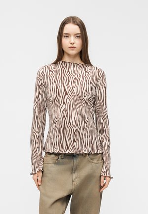 Junge Frau trägt ein langärmliges, braun-weiß gestreiftes Zebra-Top und lockere, beige Hosen, stehend vor einem schlichten weißen Hintergrund.