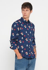 Camisa de botones azul marino con caras de Santa festivas, copos de nieve, árboles y acebo en rojo y blanco. Mangas largas con puños enrollados.