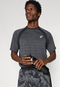 Μαύρο αθλητικό μπλουζάκι με υφή, ραγλανικές μανσέτες και λογότυπο. Το μοντέλο κρατάει μια μαύρη μπουκάλι νερού με μεταλλικό καπάκι, φορώντας σχέδια σορτς.