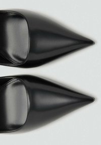 Un par de zapatos planos de cuero negro con punta afilada mostrados desde arriba sobre un fondo gris claro.