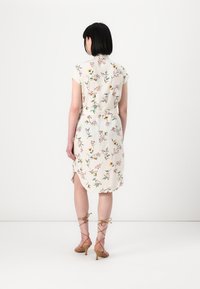 Blumenkleid aus einem weichen, hellfarbigen Stoff. Hat kurze Ärmel, eine taillierte Taille und seitliche Schlitze. Wird mit geschnürten Riemchen-Heels getragen.
