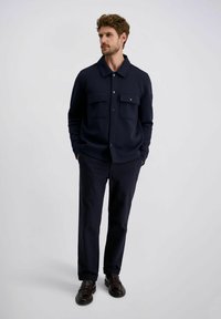  Marineblauwe shirt met lange mouwen, twee borstzakken, platte kraag en knopen. Gemaakt met donker gekleurde broeken en bruine leren schoenen.