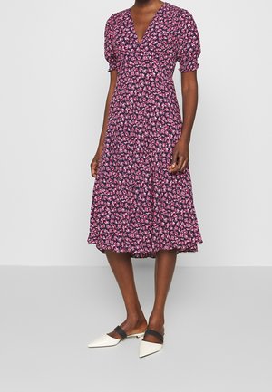 Vrouw draagt een marineblauwe jurk tot op de knie met roze bloemenprint, pofmouwen en witte puntige platte schoenen met zwarte bandjes.