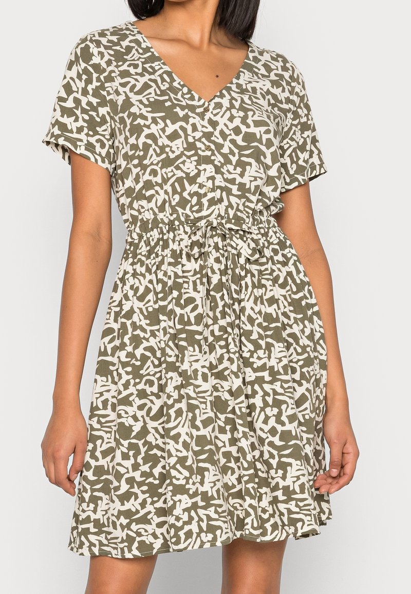 Femme portant une robe courte à manches courtes, longueur genou, avec un motif abstrait vert et blanc et une ceinture nouée à la taille.