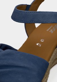 Μπλε σανδάλι από σουέντ με λουράκι στον αστράγαλο, χαρακτηριζόμενο από μαλακή υφή και σόλα από φυσικές ίνες. Εσωτερικά διαθέτει καφέ πέλμα με ενδείξεις μεγέθους.