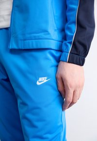 Μπλε φόρμα Nike με μανίκια που κλείνουν με φερμουάρ και παντελόνι με ελαστική μέση, φτιαγμένη από λείο ύφασμα με λευκό λογότυπο και ναυτικές λεπτομέρειες.