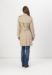 Beige trenchcoat met riem, schouderepauletten en een rugventilatie. Gedragen over blauwe flared jeans en bruine schoeisel. Minimalistisch ontwerp.