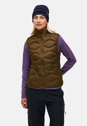 Femme portant une chemise violet à manches longues, un gilet matelassé vert olive, un pantalon noir et un bonnet rayé noir et violet.