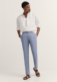 Hellblaue, maßgeschneiderte Hose mit einer strukturierten Oberfläche, kombiniert mit einem weißen Leinenhemd mit Knöpfen. Schwarze Sandalen runden den Look ab.