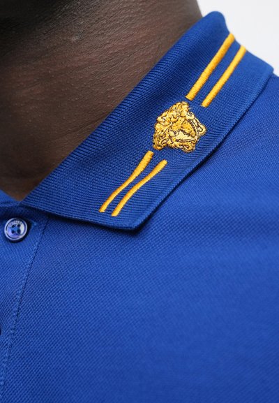 Mėlynas polo marškinėlis iš tekstūruotos medžiagos, su auksiniu liūto emblema ir trimis vertikaliomis geltonomis juostelėmis ant apykaklės. Uždarymas su dviem mygtukais.