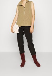 Beige hihattu ilman hihoja neulepaita, yhdistettynä mustiin capri-housuihin ja burgundinvärisiin mielekkäisiin kärkitossuihin. Pitelee pientä valkoista laukkua.