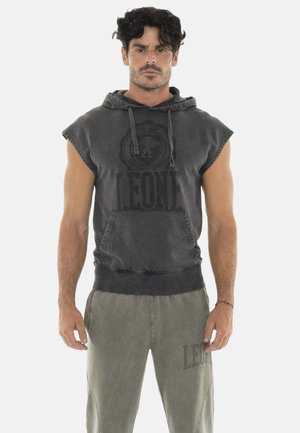 Homme aux cheveux bouclés et foncés, portant un sweat à capuche sans manches foncé et un pantalon de survêtement gris clair, debout face à un fond uni.