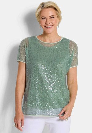 Femme souriante aux cheveux blonds courts portant un haut vert clair à manches courtes pailleté et un pantalon blanc sur un fond uni.