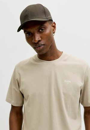 Man met een lichtbeige T-shirt met een klein wit logo en een donker olijfgroene baseballpet tegen een effen achtergrond.