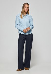 Ljusblå skjorta med knappar och långa ärmar, kombinerad med mörkblå byxor, med en subtil broderad logotyp på skjortan.