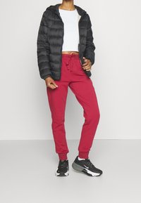 En person som bär en svart puffjacka, en vit kortärmad topp, röda joggarbyxor och svart-vita sneakers står mot en enkel bakgrund.