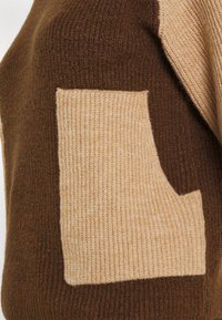 Brauner und beiger Pullover mit einem strukturierten Strickmuster und einer kontrastierenden quadratischen Brusttasche auf der linken Seite.