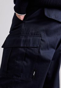 Marineblå cargobukser lavet af slidstærkt stof, med sidelommer og et lille sort logomærke på siden. Glat tekstur og skræddersyet pasform.