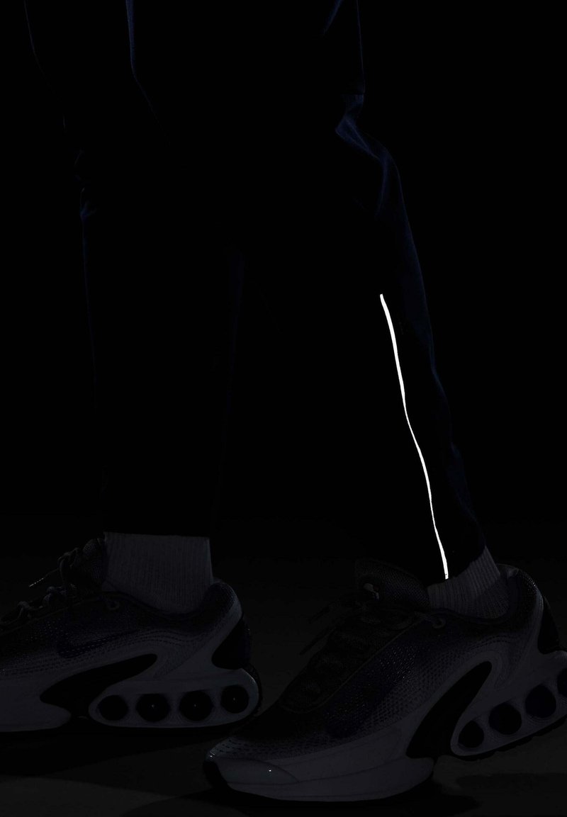 Czarne spodnie sportowe z odblaskową białą paską wzdłuż boku oraz szare sneakersy o teksturowanej górze i zaokrąglonej podeszwie.
