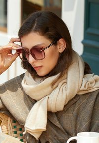 Occhiali da sole con design quadrato e lenti tintate di marrone; sciarpa beige drappeggiata sulle spalle; cappotto a zigzag in tonalità scure.