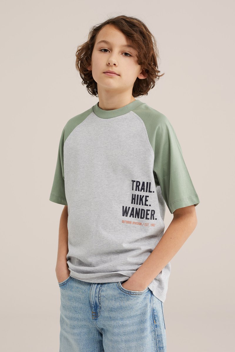 Jeune personne aux cheveux bruns ondulés portant un t-shirt gris et vert avec le texte « TRAIL. HIKE. WANDER. », mains dans les poches d'un jean bleu clair.