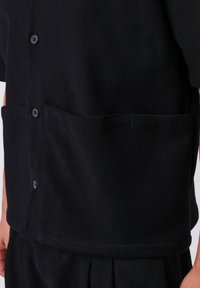 Camicia nera in tessuto strutturato con maniche corte, dotata di taschino frontale e chiusura con bottoni, che mette in evidenza un design semplice e funzionale.