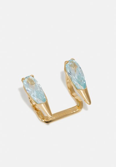 Gold ring with two elongated, light blue faceted stones at each end. Smooth, shiny finish with pointed accents. Engraved branding on the band.