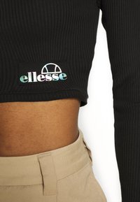 Černý žebrovaný crop top s výrazným holografickým logem „ellesse“ na náprsní kapse. V kombinaci se světle béžovými kalhotami s vysokým pasem.
