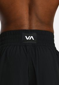 Close-up van de blote rug van een persoon die zwarte elastische shorts met een witte "VA"-logo op een patch in het midden van de taille draagt.