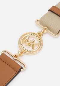 Kožený pásek v tan barvě s kruhovým logem ve zlatém tónu, které obsahuje "Michael Kors" a vzájemně propletené "MK". Béžová část z textilie s jemnou strukturou.