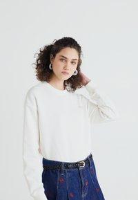 Biała bluza z długim rękawem z małym różowym logo, w połączeniu z dżinsową spódnicą z czerwonymi wzorami motyli i czarnym plecionym paskiem.