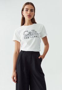 Valge puuvillane T-särk keeratud varrukatega, mille esiküljel on must kiri "Cali. THE LABEL". Selga on pandud kõrge vöökohaga mustade pükstega.
