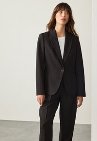 Černé sako na míru s jedním zapínacím knoflíkem, se zářezem na klopách a bočními kapsami, doplněné o úzké černé kalhoty. Hladká, strukturovaná tkanina.