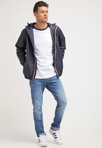 Marineblauwe hoodiejack gemaakt van zacht, waterproof materiaal met gele ritsaccenten, gedragen over een wit T-shirt en blauwe denimjeans.