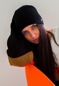 Berretto in maglia nera con una texture liscia. La persona indossa un maglione color crema. Un oggetto arancione sullo sfondo aggiunge contrasto alla scena.