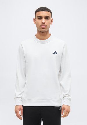 Mladý muž oblečený v bielej mikine s dlhým rukávom a malým čiernym logom na hrudi a čiernych nohaviciach, stojaci pred jednoduchým pozadím.