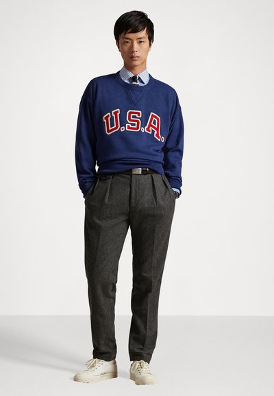 Sweatshirt marine avec "U.S.A." en lettres rouges, associé à un pantalon plissé gris. Les chaussures sont de couleur crème avec un design à lacets.
