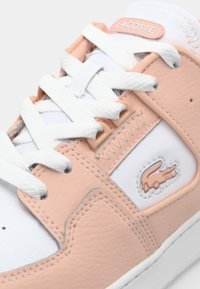 Lacoste sneakeriai su balta oda ir švelniais persikų akcentais, tekstūruotais paviršiais, baltais raišteliais ir mažu išsiuvinėtu logotipu.