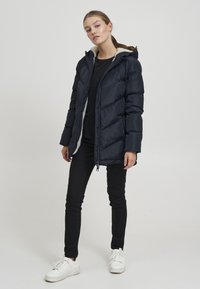 Marineblaue Daunenjacke mit strukturiertem Äußeren, gefütterter Kapuze aus Fell und Reißverschluss. Getragen über einem schwarzen Oberteil und kombiniert mit engen schwarzen Hosen und weißen Sneakern.