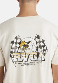 Cream cotton t-shirt featuring a large eagle graphic, checkered flags, and "RVCA" text in black and yellow at the back. Short sleeves.